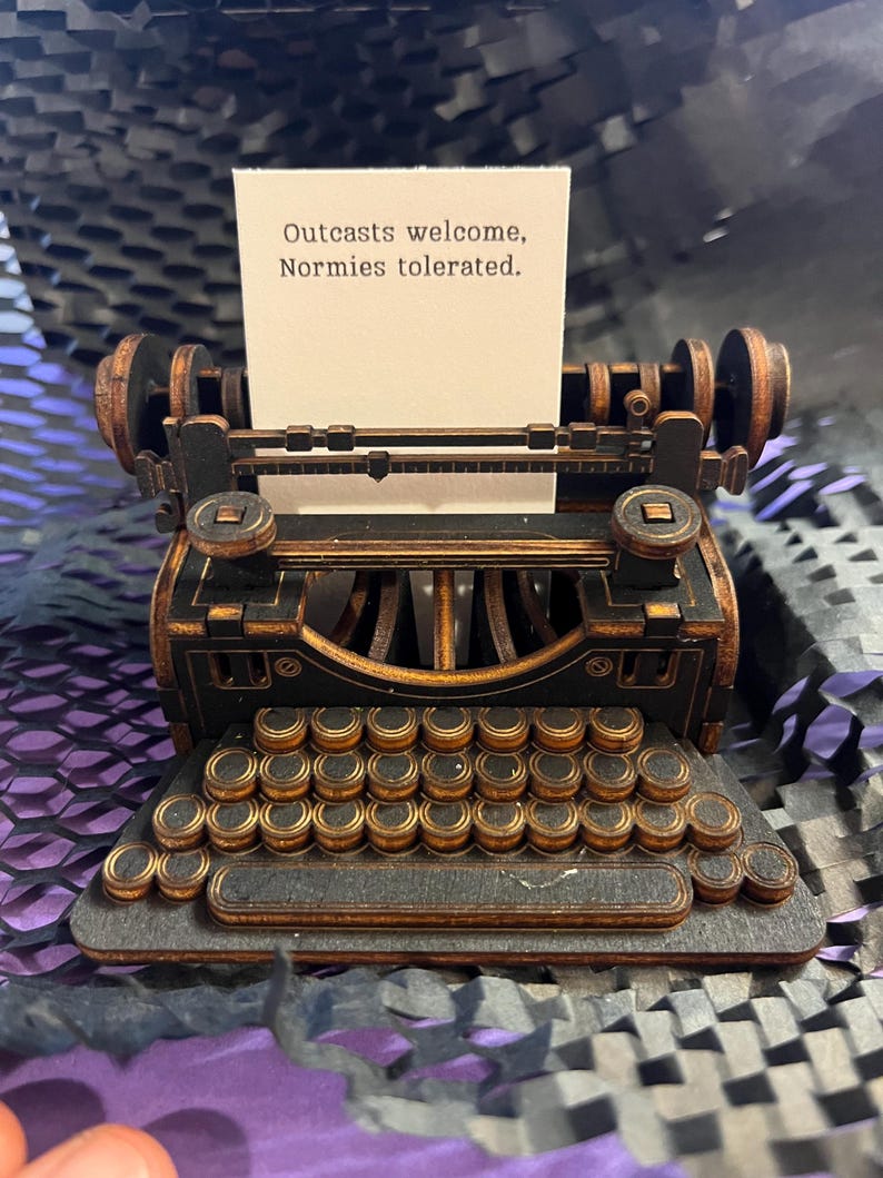 K&ouml;nnte beinhalten: Eine Miniatur-Schreibmaschine in Schwarz und Braun mit einem Papier mit der Aufschrift "Outcasts welcome, Normies tolerated." Die Schreibmaschine hat ein detailliertes Design mit Tastatur und Walzen und ist aus Holz gefertigt.