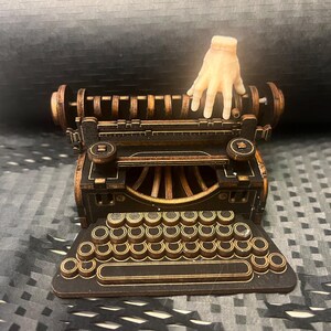 K&ouml;nnte beinhalten: Detailliertes Holzmodell einer alten Schreibmaschine mit einer Hand, die darauf ruht. Die Schreibmaschine ist haupts&auml;chlich dunkelbraun mit goldfarbenen Akzenten. Die Tastatur ist sichtbar, und das Gesamtdesign ist aufwendig.