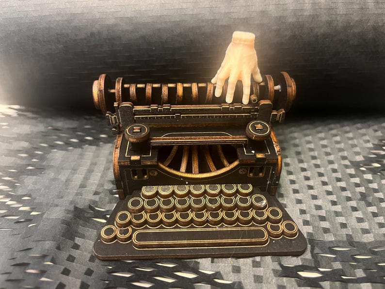K&ouml;nnte beinhalten: Detailliertes Holzmodell einer alten Schreibmaschine mit einer Hand, die darauf ruht. Die Schreibmaschine ist haupts&auml;chlich dunkelbraun mit goldfarbenen Akzenten. Die Tastatur ist sichtbar, und das Gesamtdesign ist aufwendig.