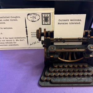 Puede incluir: Una máquina de escribir en miniatura con una estética vintage, hecha de madera oscura y metal. Cuenta con un teclado completo y detalles intrincados. Se insertan dos tarjetas, una con texto sobre pensamientos no solicitados y la otra con el texto "Outcasts welcome, Normies tolerated."