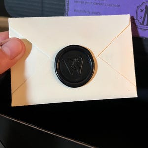 K&ouml;nnte beinhalten: Ein kleiner, cremefarbener Umschlag, der mit einem schwarzen Wachssiegel verschlossen ist. Der Stempel zeigt ein stilisiertes "W" in der Mitte. Der Umschlag ist teilweise ge&ouml;ffnet und gibt den Inhalt frei. Text auf lila Hintergrund ist sichtbar.