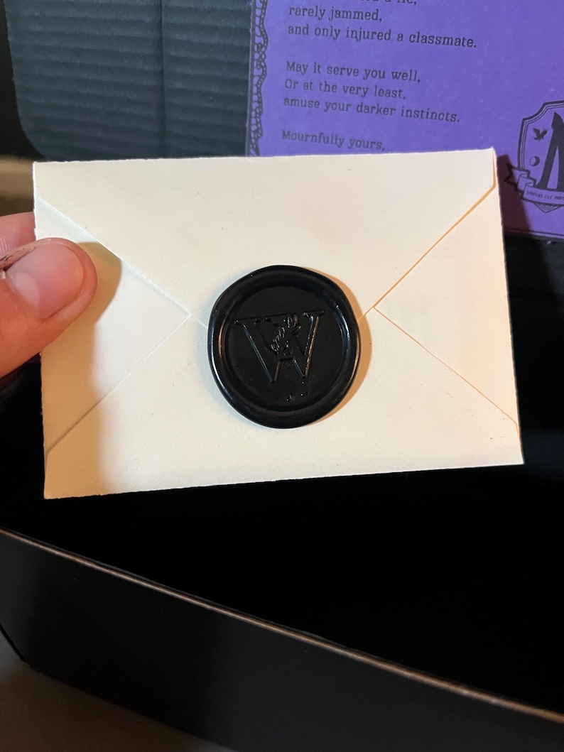 K&ouml;nnte beinhalten: Ein kleiner, cremefarbener Umschlag, der mit einem schwarzen Wachssiegel verschlossen ist. Der Stempel zeigt ein stilisiertes "W" in der Mitte. Der Umschlag ist teilweise ge&ouml;ffnet und gibt den Inhalt frei. Text auf lila Hintergrund ist sichtbar.