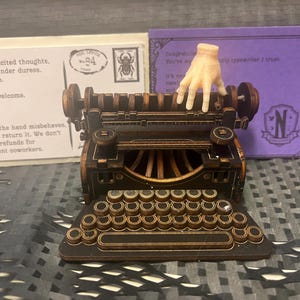 K&ouml;nnte beinhalten: Eine detaillierte Miniatur-Schreibmaschine mit Bronze- und Schwarz-Finish, mit einer vollst&auml;ndigen Tastatur und einer kleinen Handfigur. Hinter der Schreibmaschine befinden sich zwei Karten, eine mit Text und einer Briefmarke, die andere mit lila Hintergrund und dem Buchstaben "N".