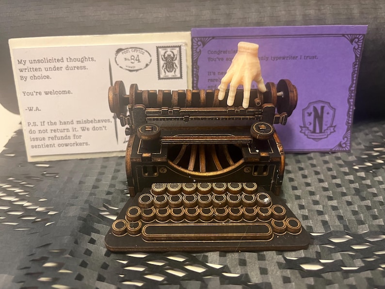 K&ouml;nnte beinhalten: Eine detaillierte Miniatur-Schreibmaschine mit Bronze- und Schwarz-Finish, mit einer vollst&auml;ndigen Tastatur und einer kleinen Handfigur. Hinter der Schreibmaschine befinden sich zwei Karten, eine mit Text und einer Briefmarke, die andere mit lila Hintergrund und dem Buchstaben "N".