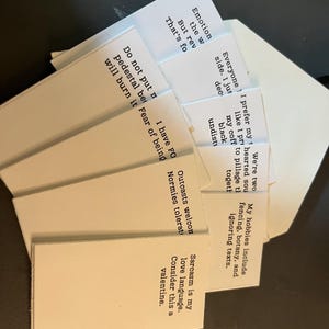 K&ouml;nnte beinhalten: Eine Sammlung wei&szlig;er rechteckiger Karten mit schwarzem Text, die auf einer dunklen Oberfl&auml;che aufgef&auml;chert sind. Die Karten enthalten verschiedene S&auml;tze und Zitate, darunter "Sarkasmus ist meine Liebessprache."