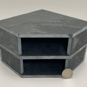 May include: A two-tiered hexagonal corner shelf made of dark gray slate. The shelf has two open compartments on each side. A US penny is placed in front of the shelf for scale.