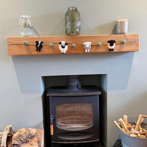 May include: A decorative garland featuring four felted sheep figures in black, white, and grey, interspersed with small sage green felt balls on a thin string. The sheep are arranged horizontally, showcasing distinct wool textures and facial details.