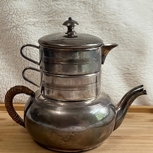 May include: A vintage silver tea set with a teapot and a separate stackable infuser. The teapot has a rounded body, a curved spout, and a woven handle. The infuser has a lid and two stacked sections.