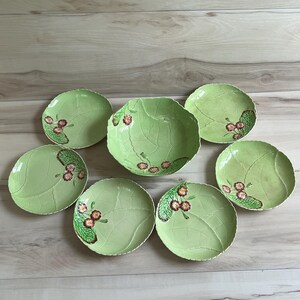 May include: A set of eight light green ceramic plates and a bowl. The plates and bowl are leaf-shaped with detailed veining and a floral design in shades of red, brown, and green. The set is arranged on a light wood surface.