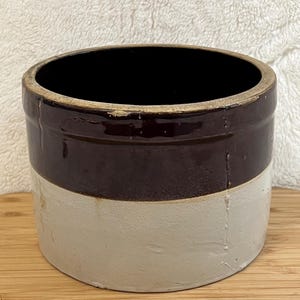 May include: A two-tone stoneware crock with a dark brown upper section and a cream-colored lower section. The antique container has a wide opening and a simple, classic design, suitable for kitchen or home decor.