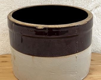 May include: A two-tone stoneware crock with a dark brown upper section and a cream-colored lower section. The antique container has a wide opening and a simple, classic design, suitable for kitchen or home decor.