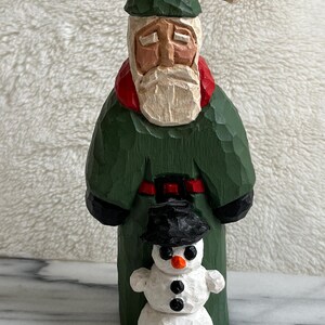 May include: Hand-carved wooden Santa Claus figure in a green coat, red collar, and black belt, with a small snowman in front. The snowman has a black hat, coal eyes, and a carrot nose. The figures are on a white surface.