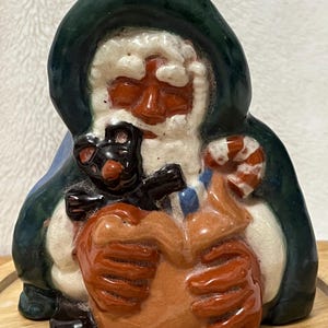 May include: A ceramic Santa Claus figurine with a green hood, white beard, and red face. He holds a black cat and a candy cane, with a brown sack. The figurine has a glossy finish and is likely a Christmas decoration.