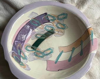 Może przedstawiać: Duża, okrągła ceramiczna miska z abstrakcyjnym wzorem w odcieniach fioletu, różu, zieleni i żółci. Miska ma jasnofioletowy rant i białe wnętrze. Podpis artysty, "Gilmore", jest widoczny na spodzie miski.