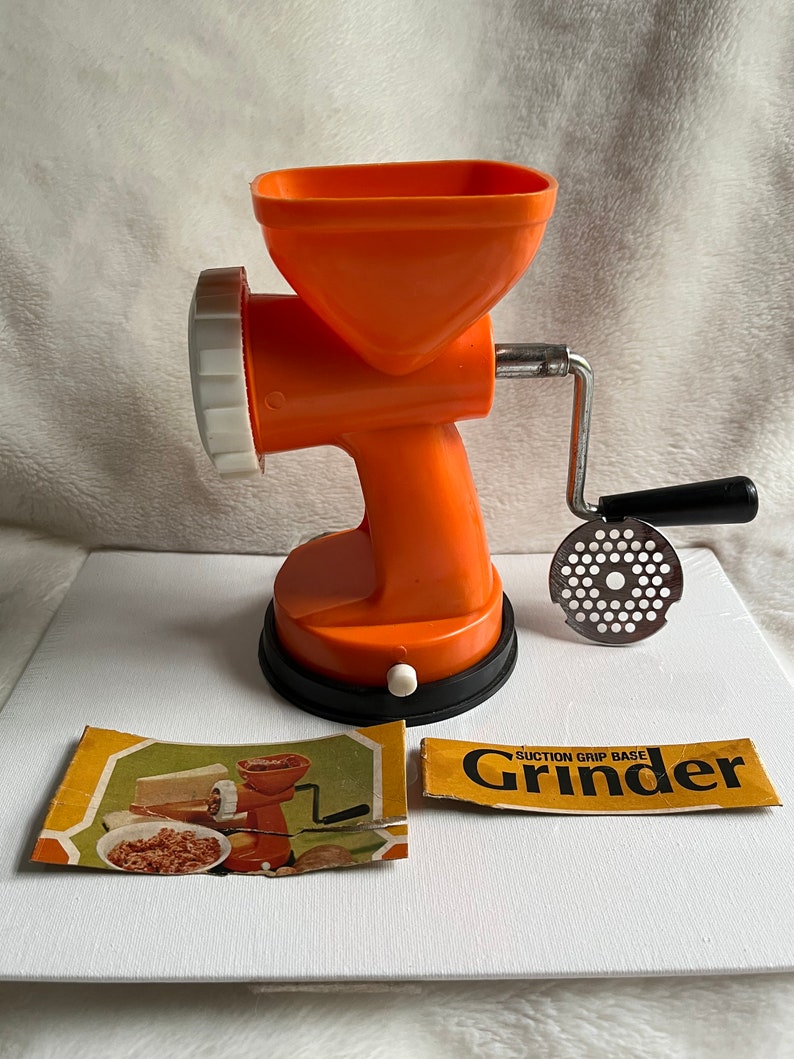 A vintage handheld meat grinder with an orange body, a black handle, and a metal grinding disc, accompanied by instruction leaflets and a "Suction Grip Base" label.