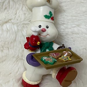 May include: A snowman Christmas ornament wearing a chef's hat and holding a tray of cookies. The snowman has a red bird on its arm and is wearing red boots. The ornament is white, red, green, and brown.