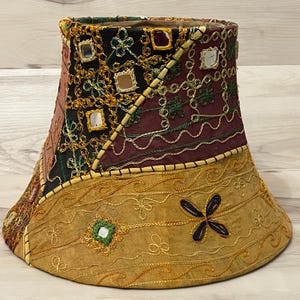 May include: A decorative lampshade with a patchwork design. The shade features a mix of colors including gold, burgundy, and black, with embroidered details and small mirror accents. The base is wider than the top.