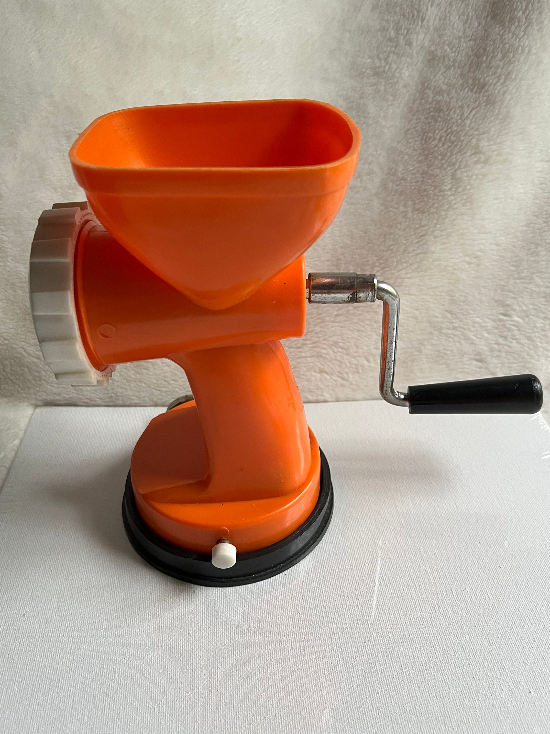 A vintage orange manual meat grinder with a black base is displayed on a white surface.