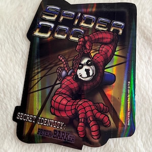 May include: A holographic sticker featuring a cartoon dog dressed as Spider-Man. The sticker has a black background with a silver and gold iridescent sheen. The text "Spider Dog" is at the top of the sticker and the text "Secret Identity: Peter Barker" is at the bottom. The text "& 2004 Big Dog Sportswear. This is a Parody" is in the bottom right corner.