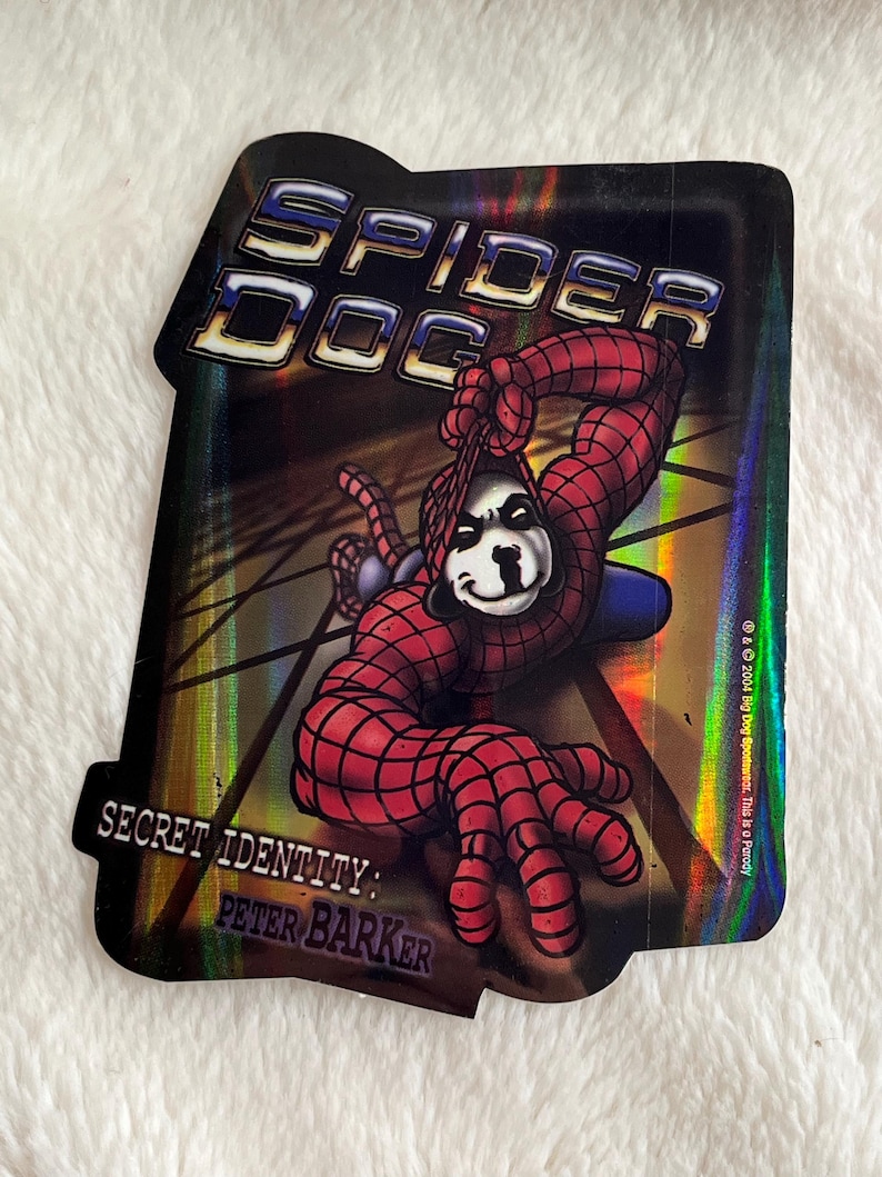 May include: A holographic sticker featuring a cartoon dog dressed as Spider-Man. The sticker has a black background with a silver and gold iridescent sheen. The text "Spider Dog" is at the top of the sticker and the text "Secret Identity: Peter Barker" is at the bottom. The text "& 2004 Big Dog Sportswear. This is a Parody" is in the bottom right corner.