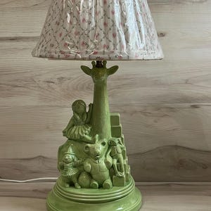 May include: A table lamp with a light green ceramic base featuring sculpted figures of a giraffe, a doll, a teddy bear, a monkey, and stacked blocks. The lamp is topped with a conical fabric lampshade decorated with a small pink floral pattern.