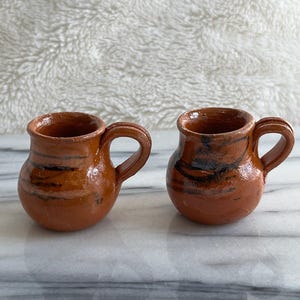 May include: Two small, handmade, brown ceramic mugs with rounded bodies and curved handles. Each mug has dark brown and black streaks and a glossy finish. The mugs are displayed on a marble surface.
