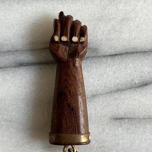May include: A carved wooden fist bottle stopper. The fist is brown with white accents on the knuckles. The handle is a dark brown with a brass band and a small metal loop at the bottom. The stopper is designed to fit into a bottle.