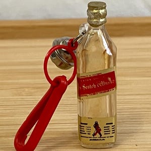 Puede incluir: Llavero de botella de vidrio transparente en miniatura con una etiqueta roja que dice "Scotch Whisky". La botella tiene una tapa dorada y un clip rojo. La etiqueta presenta una figura caminando y los a&ntilde;os 1865, 1867 y 1887.