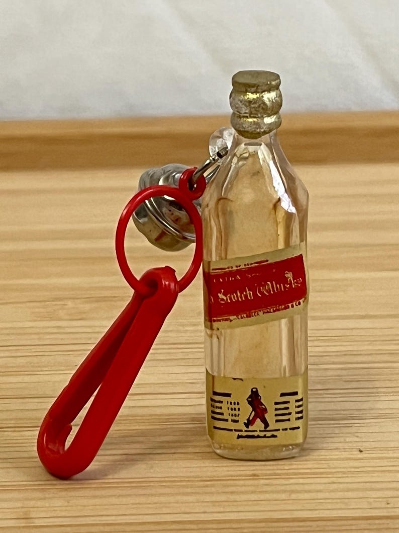 Puede incluir: Llavero de botella de vidrio transparente en miniatura con una etiqueta roja que dice "Scotch Whisky". La botella tiene una tapa dorada y un clip rojo. La etiqueta presenta una figura caminando y los a&ntilde;os 1865, 1867 y 1887.