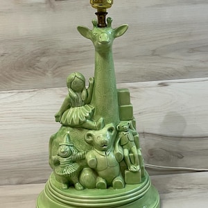 May include: A light green ceramic table lamp base featuring a tall giraffe neck and a collection of molded toys including a doll, a teddy bear, a clown, and stacked blocks. The lamp has a gold-colored metal socket at the top for a light bulb.