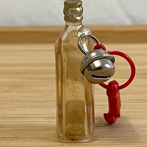 Puede incluir: Una peque&ntilde;a botella de vidrio transparente con una tapa dorada. Se adjunta una campana plateada y un dije de pl&aacute;stico rojo. La botella est&aacute; sobre una superficie de madera marr&oacute;n claro.