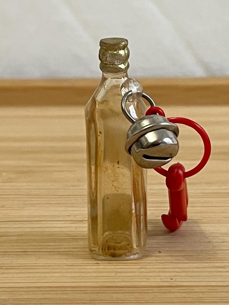 Puede incluir: Una peque&ntilde;a botella de vidrio transparente con una tapa dorada. Se adjunta una campana plateada y un dije de pl&aacute;stico rojo. La botella est&aacute; sobre una superficie de madera marr&oacute;n claro.
