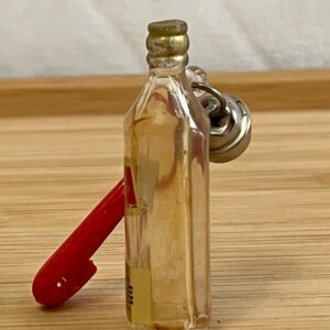 Puede incluir: Una peque&ntilde;a botella de vidrio con tapa dorada y un llavero de metal plateado. Un bol&iacute;grafo rojo se apoya en la botella. La botella tiene una etiqueta amarilla con texto.