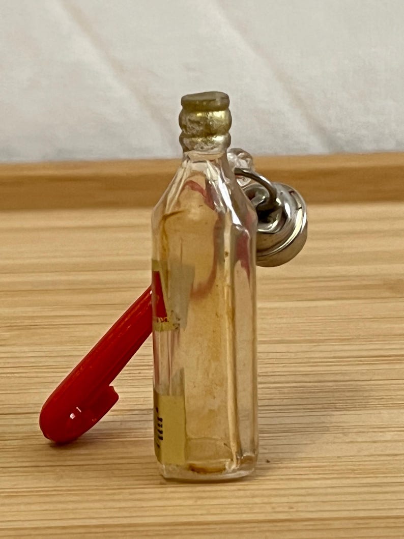 Puede incluir: Una peque&ntilde;a botella de vidrio con tapa dorada y un llavero de metal plateado. Un bol&iacute;grafo rojo se apoya en la botella. La botella tiene una etiqueta amarilla con texto.