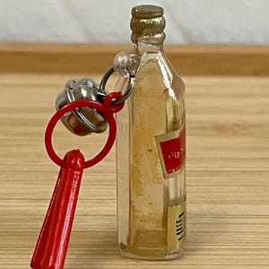 Puede incluir: Botella peque&ntilde;a de vidrio transparente con tapa dorada y etiqueta roja. La botella est&aacute; unida a un llavero de pl&aacute;stico rojo con una campana plateada. El llavero est&aacute; sobre una superficie de madera.