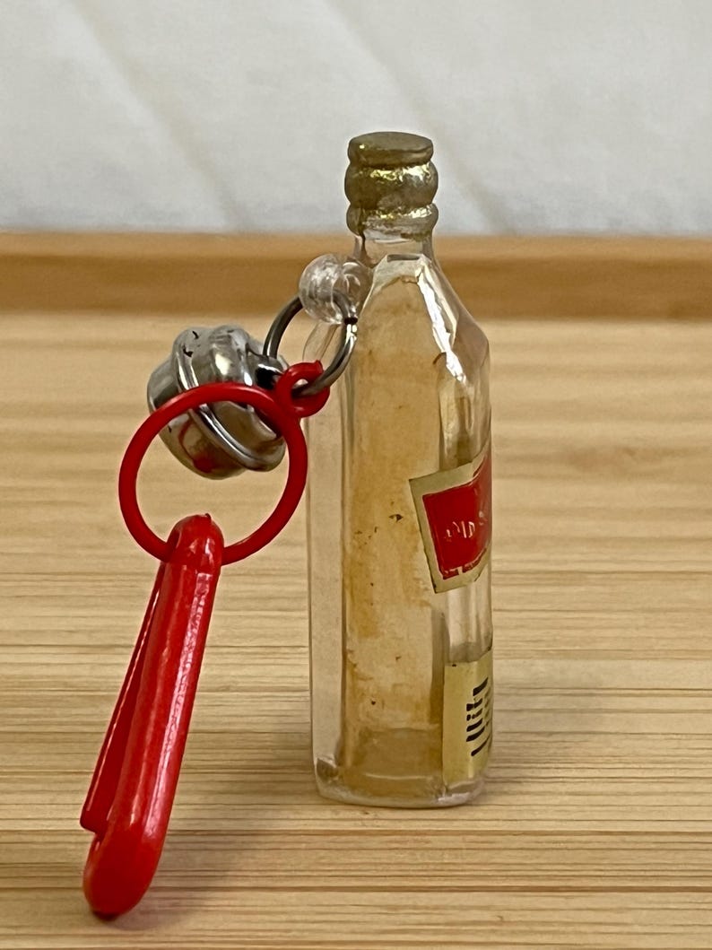 Puede incluir: Botella peque&ntilde;a de vidrio transparente con tapa dorada y etiqueta roja. La botella est&aacute; unida a un llavero de pl&aacute;stico rojo con una campana plateada. El llavero est&aacute; sobre una superficie de madera.