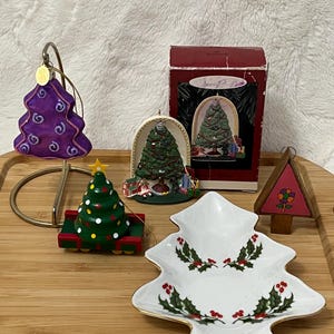 May include: A collection of Christmas ornaments and a tree-shaped dish. The ornaments include a purple tree, a green tree on a red base, and a wooden triangle with a flower. The dish is white with a gold rim and holly berry design.
