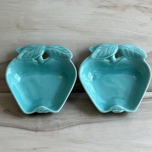 May include: Two turquoise ceramic apple-shaped dishes. Each dish has a leaf design at the top and a small hole. The dishes are a bright, solid color and are placed side by side.