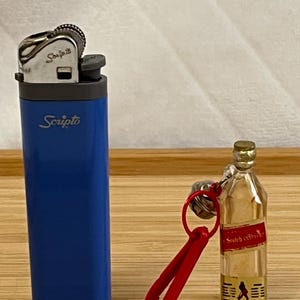 Puede incluir: Un encendedor Scripto azul y una botella en miniatura de whisky escoc&eacute;s con un llavero rojo. El encendedor tiene el logotipo de Scripto en blanco. La botella en miniatura tiene una etiqueta roja y un logotipo de un hombre caminando.