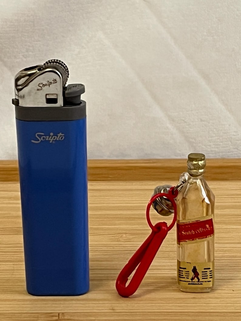Puede incluir: Un encendedor Scripto azul y una botella en miniatura de whisky escoc&eacute;s con un llavero rojo. El encendedor tiene el logotipo de Scripto en blanco. La botella en miniatura tiene una etiqueta roja y un logotipo de un hombre caminando.