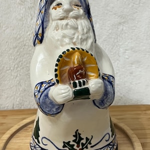 May include: A ceramic Santa Claus figurine, predominantly white with blue and yellow accents. The figure holds a candle holder with a lit candle. The base features green holly and a decorative pattern. A festive Christmas decoration.