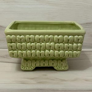 May include: A rectangular light green ceramic planter with a raised, repeating U-shaped texture on the exterior walls. The planter features a wide, open top and is supported by a sturdy, integrated base with matching textured detailing on the sides.