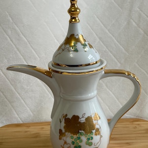 May include: White ceramic coffee pot with gold trim and a gold finial. The pot features a decorative design of gold grape leaves and green grapes. The handle and spout are also trimmed in gold. The pot is sitting on a wooden surface.