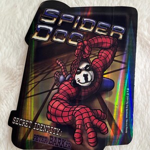 May include: A holographic sticker featuring a cartoon dog dressed as Spider-Man. The sticker has the text "Spider Dog" and "Secret Identity: Peter Barker".