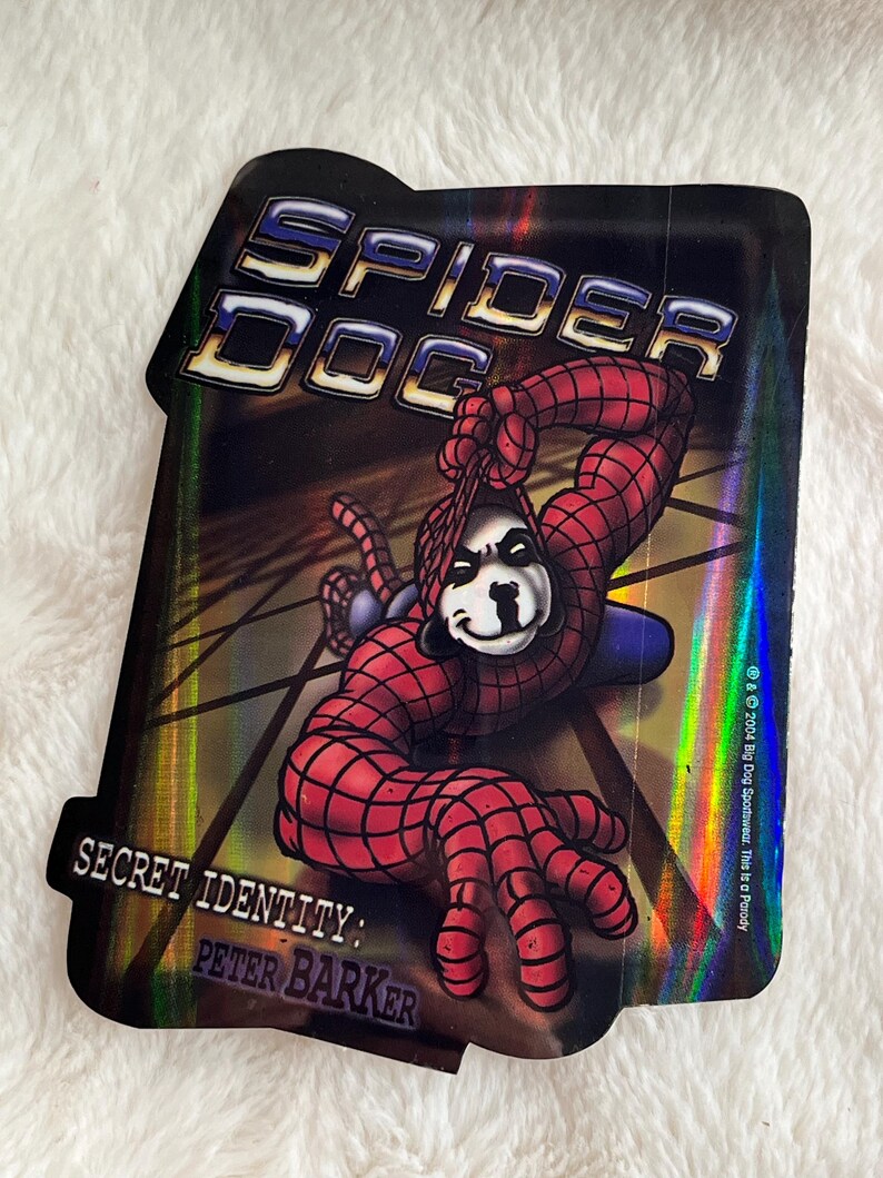 May include: A holographic sticker featuring a cartoon dog dressed as Spider-Man. The sticker has the text "Spider Dog" and "Secret Identity: Peter Barker".