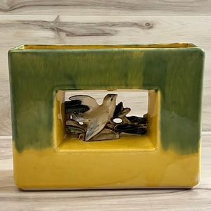 May include: A rectangular ceramic vase featuring a two-tone glaze with a vibrant green top and a bright yellow base. The center of the vase contains a cut-out window displaying a sculpted ceramic bird perched on a branch with dark accents.