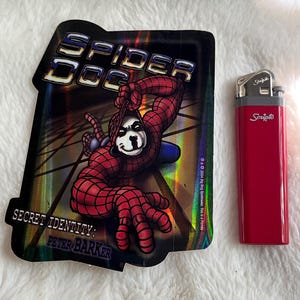 May include: A holographic sticker featuring a cartoon dog dressed as Spider-Man. The sticker has the text "Spider Dog" and "Secret Identity: Peter Barker".