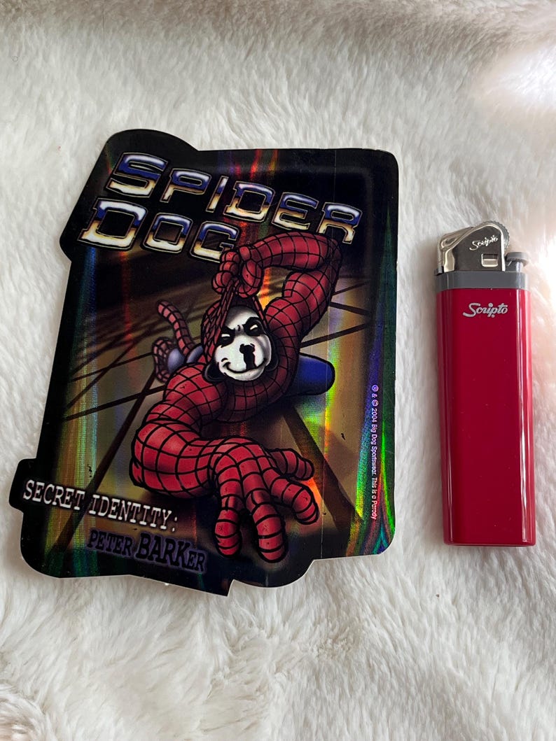 May include: A holographic sticker featuring a cartoon dog dressed as Spider-Man. The sticker has the text "Spider Dog" and "Secret Identity: Peter Barker".