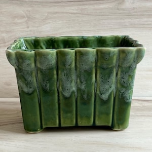 May include: A rectangular, green ceramic planter with a ribbed design. The planter has a glossy finish with lighter green accents along the top edges. The planter is a decorative item for indoor or outdoor use.