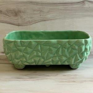 May include: A rectangular, light green ceramic planter with a textured, geometric pattern. The planter sits on three small, rounded feet. The interior is smooth, and the exterior features a repeating, raised design. This decorative piece is suitable for indoor or outdoor use.